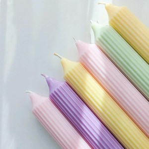 May include: A collection of pastel-colored, ribbed candles. The candles are arranged diagonally, showcasing a palette of soft hues including pink, yellow, green, and purple. Each candle has a pointed tip with a visible wick, suggesting they are ready for use.