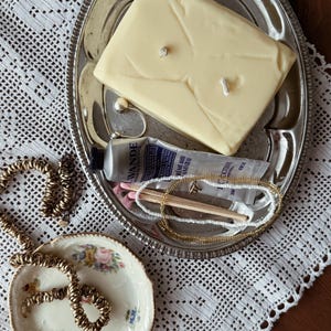 May include: A rectangular cream-coloured candle with two wicks, a silver-toned metal tray, a tube of L'Occitane hand cream, a small pile of matches, a gold-toned chain necklace, and a small floral-patterned ceramic dish with a gold chain.