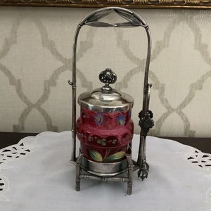 Puede incluir: Un soporte de servicio plateado con un tarro de vidrio con un diseño floral rojo y blanco. El soporte tiene un diseño de estilo bambú y el tarro tiene una tapa plateada.