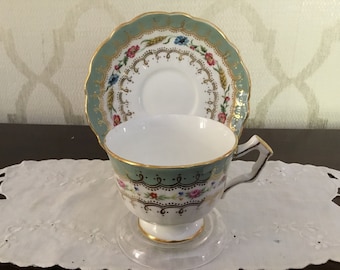 May include: A delicate teacup and saucer set with a green and gold floral pattern. The cup and saucer are both trimmed with gold accents and feature a floral design with pink, blue, and yellow flowers.
