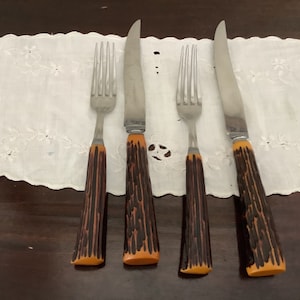 Peut inclure: Ensemble de couverts vintage de quatre pièces, comprenant deux fourchettes et deux couteaux. Les lames en acier inoxydable sont associées à des manches avec un motif imitation bois de cerf marron et orange. L'ensemble est disposé sur un tissu blanc brodé.