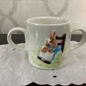 以下が含まれることがあります： 2つの取っ手がついた白いセラミック製のマグカップ。3Dの画像で、赤ちゃんウサギを抱いた母親ウサギが描かれています。ウサギは青いドレスを着て、木の椅子に座っています。