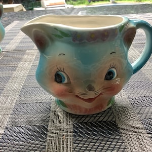 Peut inclure: Une crémier en céramique bleue avec un visage de chat de dessin animé. Le chat a des joues roses, de grands yeux bleus et une bouche rose. La crémier a une poignée blanche et un motif floral autour du haut.