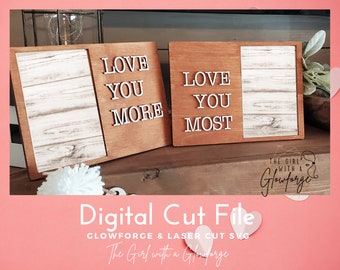 May include: Two wooden signs with a white woodgrain background. The signs have the text "LOVE YOU MORE" and "LOVE YOU MOST" in white letters.