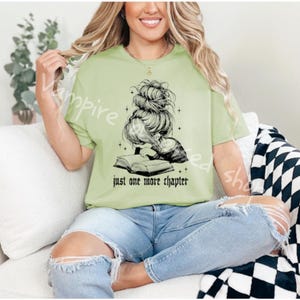 May include: A light green t-shirt with the text "just one more chapter" and a black graphic of a woman reading a book. The woman is wearing her hair in a messy bun. The person in the image is wearing blue jeans.