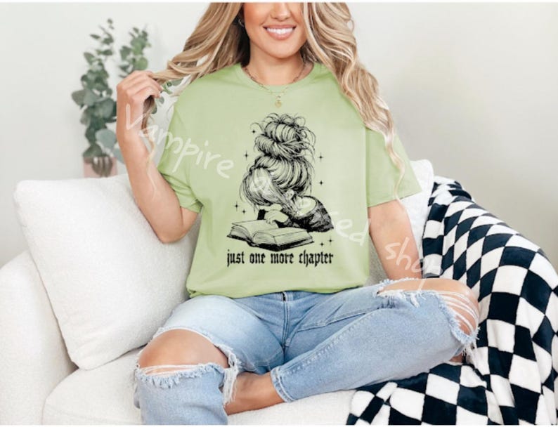 May include: A light green t-shirt with the text "just one more chapter" and a black graphic of a woman reading a book. The woman is wearing her hair in a messy bun. The person in the image is wearing blue jeans.