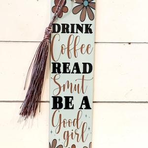 May include: A light blue bookmark with a tassel. The bookmark features the words "DRINK Coffee READ Smut BE A Good girl" in black and brown text. It is decorated with brown and blue flowers.