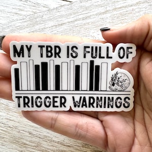 May include: White sticker with black text that reads "My TBR is full of" and a barcode graphic. A small black and white skull with leaves is on the right side of the barcode. The text "Trigger Warnings" is below the barcode.