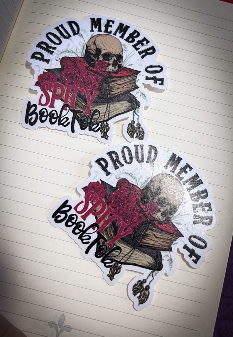 May include: Two white stickers with black text that reads "Proud Member of Spicy BookTok". Each sticker features a skull with a red and black book stack and floral accents.