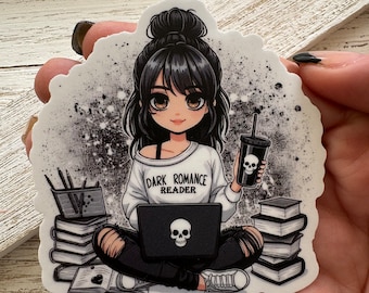Puede incluir: Una pegatina con una ilustración de estilo anime de una persona con cabello negro, que lleva un suéter que dice "Dark Romance Reader". La persona está sentada con una computadora portátil y una bebida adornada con una calavera. Los libros rodean a la persona.