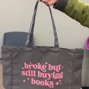 Op de afbeelding: Grijze draagtas met de roze tekst "broke but still buying books". De tas heeft lange handvatten en wordt door een persoon omhoog gehouden. De achtergrond is een effen muur.