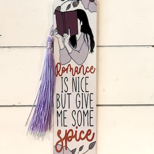 May include: A white bookmark with a lilac tassel. The bookmark features an illustration of a person reading a book, with the text "Romance is nice but give me some spice". Floral accents are also included.