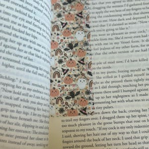 May include: A close-up of a book with a Halloween-themed bookmark. The bookmark features a variety of spooky and cute illustrations, including pumpkins, ghosts, bats, and witches. The text on the bookmark reads "The Cursed".