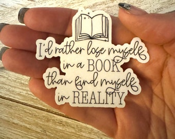 Puede incluir: Pegatina blanca con texto negro y un dibujo de un libro abierto con estrellas. El texto dice "Prefiero perderme en un libro que encontrarme en la realidad."