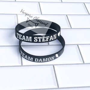 May include: Two black silicone bracelets with white text. The top bracelet says "Team Stefan" with a heart symbol. The bottom bracelet says "Team Damon" with a bird symbol.