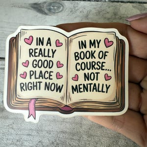 May include: A sticker in the shape of an open book with the text "IN A REALLY GOOD PLACE RIGHT NOW" on the left page and "IN MY BOOK OF COURSE... NOT MENTALLY" on the right page. Pink hearts are scattered throughout the text.
