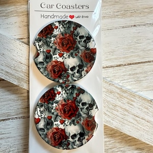 May include: Two car coasters with a design of skulls, red roses, and hearts. The coasters are round and have a small indentation for a car cup holder. The packaging reads "Car Coasters Handmade with love."