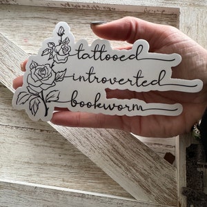 May include: White sticker with a black line drawing of a rose and the text "tattooed introvert bookworm".