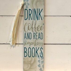 May include: A rectangular bookmark with the text "DRINK Coffee AND READ romantasay BOOKS" in teal and grey lettering. The bookmark has a floral design and a cream-coloured tassel. The background is a white wooden surface.