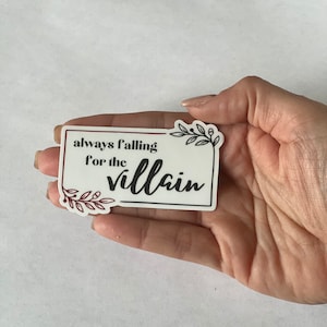 May include: A white sticker with a burgundy border and black text that reads "always falling for the villain". The sticker has a floral design in burgundy.