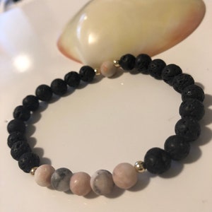 May include: A bracelet made of black lava stone and gemstones. The bracelet has black, round lava beads and pink and grey gemstone beads. Gold-coloured beads separate the different bead types. The bracelet is on a white surface.