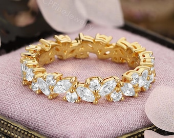 May include: A gold-colored ring with a continuous pattern of marquise-cut white gemstones. The ring is displayed on a small, ornate box with a pink interior, surrounded by soft pink and white floral accents. The ring's design is elegant and eye-catching.
