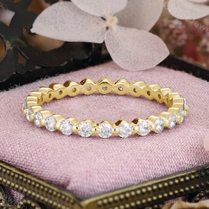 May include: A gold eternity ring with a continuous row of round, clear gemstones. The ring is set on a pink velvet surface inside a decorative, antique-style box. The box has a dark, ornate design.