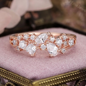 May include: Rose gold ring featuring a cluster of various-shaped clear gemstones. The ring's design includes round, pear, and square-cut stones, creating a sparkling, asymmetrical arrangement. The ring is set on a pink velvet surface.