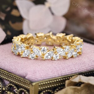May include: A gold-colored ring with a continuous pattern of marquise-cut white gemstones. The ring is set on a pink surface inside a decorative box. The background features blurred floral elements and the text "CPFineJewelry".