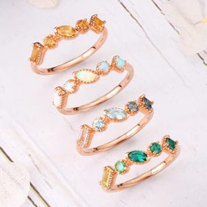 May include: Four rose gold rings with colorful gemstones arranged in a row. The gemstones include yellow, orange, white, blue, and green. The rings are displayed on a white wooden surface.