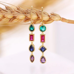 May include: A pair of gold-colored drop earrings with a series of colorful gemstones. Each earring features a teardrop, rectangle, and diamond-shaped stone in shades of green, pink, blue, and purple. The earrings are suspended from a gold hook.
