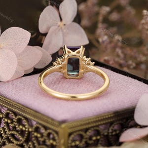 Puede incluir: Un anillo de oro con una piedra preciosa rectangular de color verde azulado y piedras de acento m&aacute;s peque&ntilde;as a cada lado. El anillo est&aacute; sobre una superficie rosa, con las palabras "CPFineJewelry" visibles en el fondo.