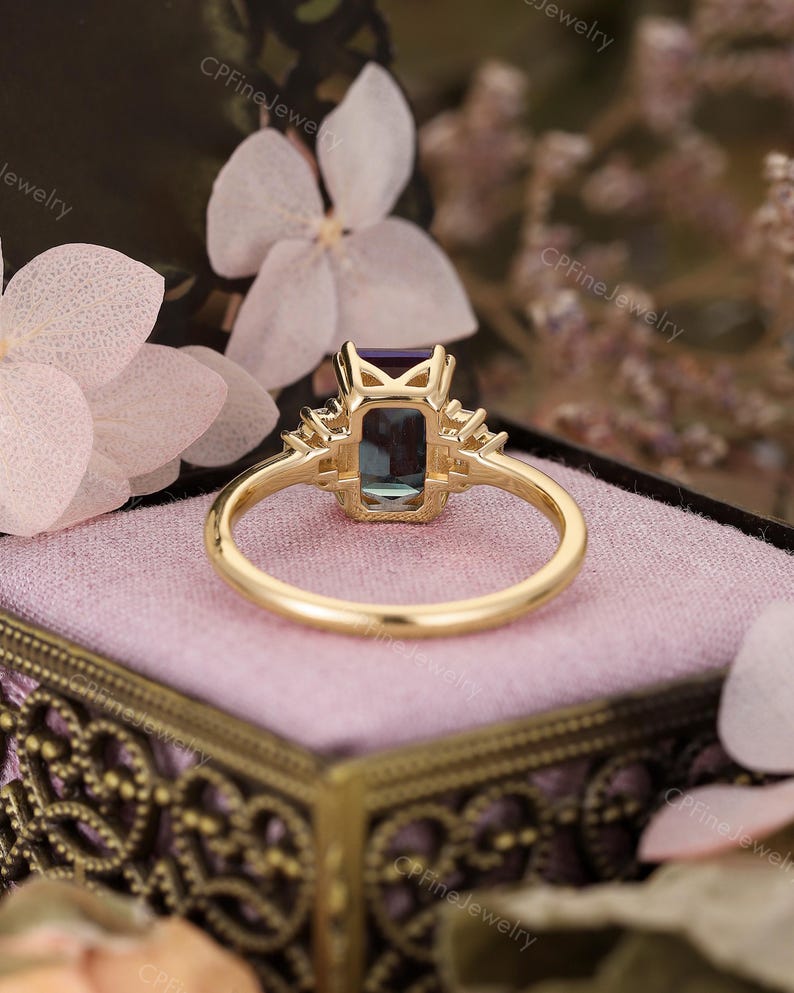Puede incluir: Un anillo de oro con una piedra preciosa rectangular de color verde azulado y piedras de acento m&aacute;s peque&ntilde;as a cada lado. El anillo est&aacute; sobre una superficie rosa, con las palabras "CPFineJewelry" visibles en el fondo.