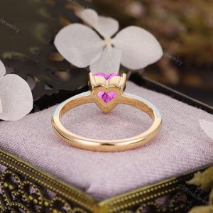 May include: A gold ring featuring a heart-shaped pink gemstone set in a prong setting. The band is accented with a smooth white enamel inlay along the top half, creating a contrast against the polished gold metal finish.