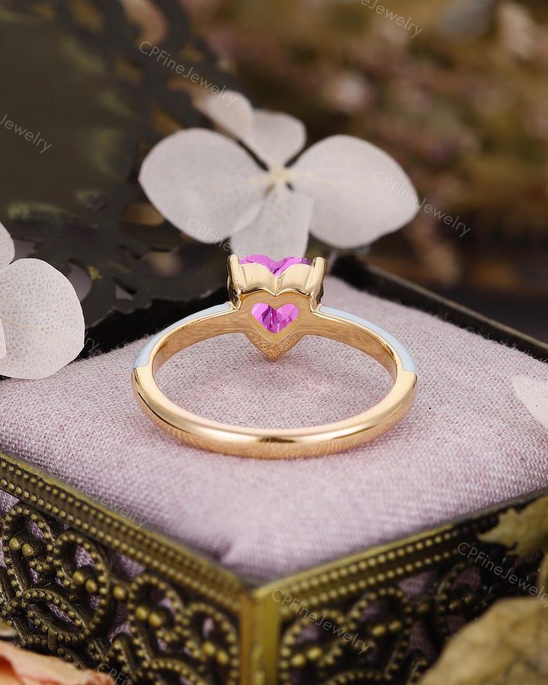 May include: A gold ring featuring a heart-shaped pink gemstone set in a prong setting. The band is accented with a smooth white enamel inlay along the top half, creating a contrast against the polished gold metal finish.