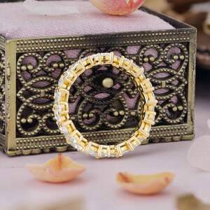 May include: A gold eternity ring with square-cut, clear gemstones. The ring is set in a gold band and is displayed on a pink velvet surface inside a decorative box. Scattered petals add a delicate touch.