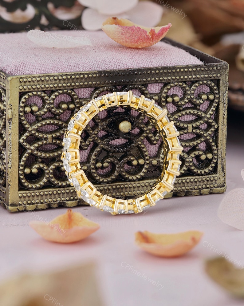 May include: A gold eternity ring with square-cut, clear gemstones. The ring is set in a gold band and is displayed on a pink velvet surface inside a decorative box. Scattered petals add a delicate touch.