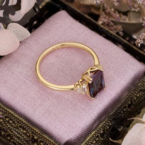 Puede incluir: Un anillo de oro con una piedra preciosa rectangular de color p&uacute;rpura oscuro. El anillo tiene una banda delicada y est&aacute; engastado con piedras m&aacute;s peque&ntilde;as y claras a ambos lados de la piedra principal. El anillo se muestra en una caja decorativa con interior rosa.