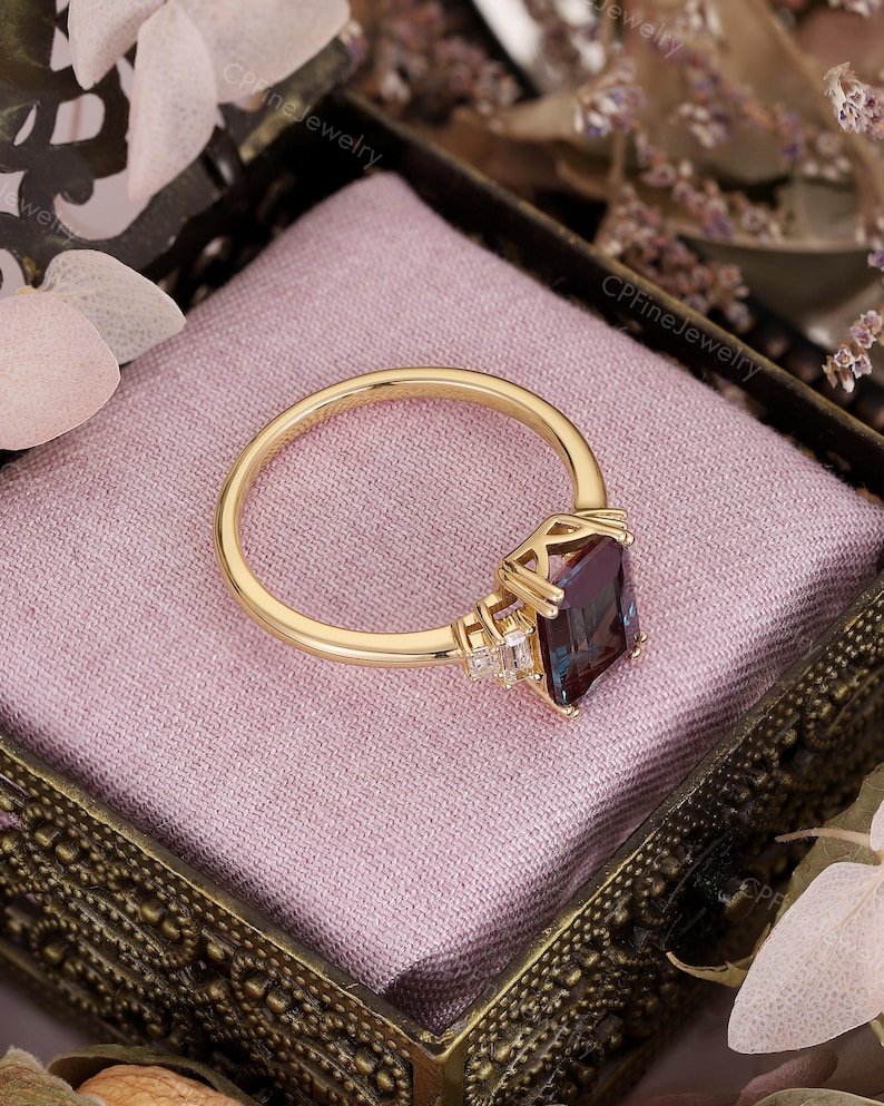 Puede incluir: Un anillo de oro con una piedra preciosa rectangular de color p&uacute;rpura oscuro. El anillo tiene una banda delicada y est&aacute; engastado con piedras m&aacute;s peque&ntilde;as y claras a ambos lados de la piedra principal. El anillo se muestra en una caja decorativa con interior rosa.