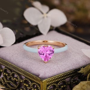 May include: A ring featuring a heart-shaped pink gemstone set in a rose gold band with a light blue enamel finish. The ring is displayed on a soft, light-colored cushion inside a decorative box with ornate metal detailing.