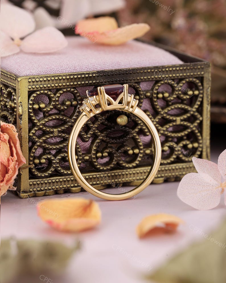 Puede incluir: Un anillo de oro con una gema rectangular de color rojo oscuro como pieza central. El anillo tiene gemas m&aacute;s peque&ntilde;as y claras a ambos lados de la piedra principal. El anillo se encuentra sobre un joyero decorativo de estilo antiguo y con detalles florales.