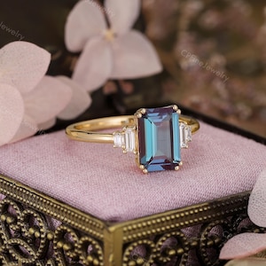Puede incluir: Un anillo de oro con una piedra preciosa rectangular de color turquesa como pieza central, flanqueada por piedras preciosas blancas rectangulares m&aacute;s peque&ntilde;as. El anillo est&aacute; sobre una superficie rosa, con detalles florales en el fondo.