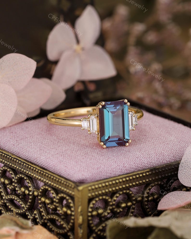Puede incluir: Un anillo de oro con una piedra preciosa rectangular de color turquesa como pieza central, flanqueada por piedras preciosas blancas rectangulares m&aacute;s peque&ntilde;as. El anillo est&aacute; sobre una superficie rosa, con detalles florales en el fondo.