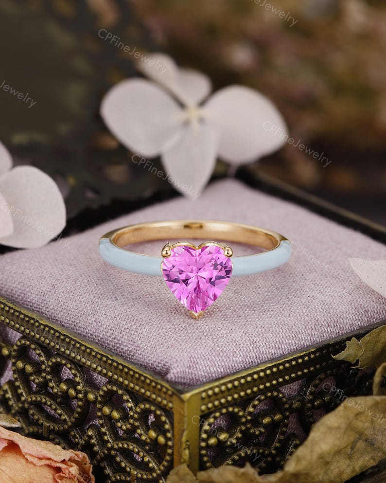 May include: A gold ring featuring a heart-shaped pink gemstone set in the center. The band of the ring is finished with a smooth, light blue enamel coating. The ring is positioned on a soft, light purple fabric surface.