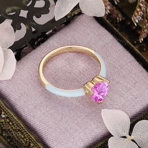 May include: A gold ring featuring a heart-shaped pink gemstone set in a prong setting. The band is partially coated in a smooth, light blue enamel finish, creating a contrast between the metallic gold, the vibrant pink stone, and the blue.