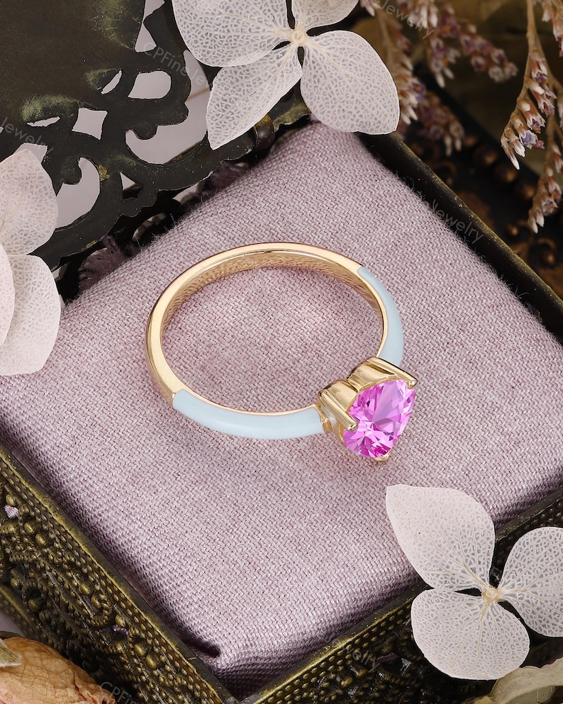 May include: A gold ring featuring a heart-shaped pink gemstone set in a prong setting. The band is partially coated in a smooth, light blue enamel finish, creating a contrast between the metallic gold, the vibrant pink stone, and the blue.