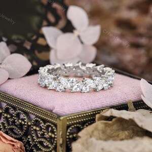 May include: A silver ring with a continuous pattern of marquise-cut white gemstones. The ring is set on a pink velvet surface inside a decorative box. The background features soft focus flowers and foliage.