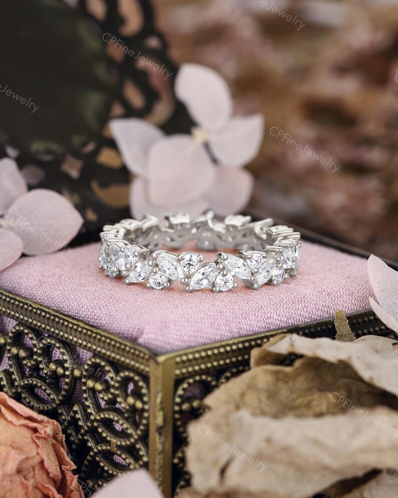 May include: A silver ring with a continuous pattern of marquise-cut white gemstones. The ring is set on a pink velvet surface inside a decorative box. The background features soft focus flowers and foliage.
