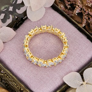 May include: A gold-colored eternity ring with clear, rectangular-cut stones set around the band. The ring is displayed in a decorative box with a pink interior, surrounded by floral accents.