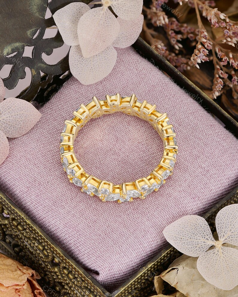 May include: A gold-colored eternity ring with clear, rectangular-cut stones set around the band. The ring is displayed in a decorative box with a pink interior, surrounded by floral accents.